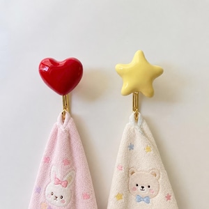 以下が含まれることがあります： ゴールドトーンの金属製ベースが付いた2つの壁掛けフック。1つは光沢のある赤いハート、もう1つは光沢のある淡い黄色の星が上部に付いています。各フックには、ウサギのグラフィックが描かれたピンクのタオルと、クマのグラフィックが描かれたクリーム色のタオルが掛けられています。