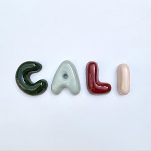 May include: Four ceramic letters spell "CALI". The 'C' is dark green, the 'A' is light grey, the 'L' is burgundy, and the 'I' is pale pink. The letters are arranged horizontally against a white background, with a green border.