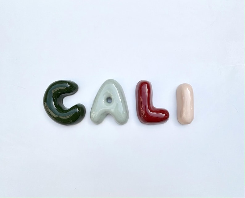 May include: Four ceramic letters spell "CALI". The 'C' is dark green, the 'A' is light grey, the 'L' is burgundy, and the 'I' is pale pink. The letters are arranged horizontally against a white background, with a green border.