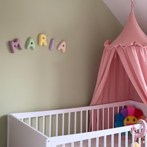 May include: A nursery scene featuring a white crib, a pink canopy, and the name "MARIA" spelled out in colorful, 3D letters on the wall. A multi-colored flower-shaped pillow is visible inside the crib.