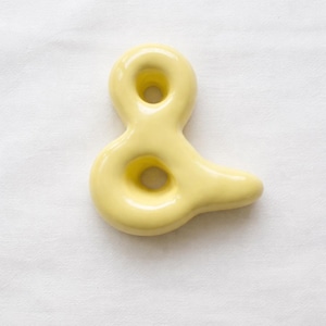 May include: A pale yellow ceramic ampersand sculpture. The ampersand has a smooth, glossy finish and a rounded shape with two circular openings. The sculpture is set against a white background.