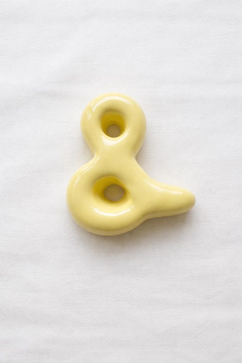 May include: A pale yellow ceramic ampersand sculpture. The ampersand has a smooth, glossy finish and a rounded shape with two circular openings. The sculpture is set against a white background.