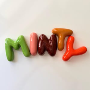 Op de afbeelding: Een set van vijf glanzende, afgeronde, driedimensionale letters die het woord MINTY vormen. De letters zijn gekleurd in limoengroen, lichtroze, donkerbruin, mosterdgeel en feloranje, elk met een gladde, reflecterende oppervlakteafwerking.