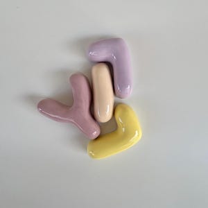 May include: Four abstract ceramic shapes in pastel colours: lavender, yellow, pink, and beige. The shapes are smooth and glossy, with rounded edges. The arrangement is scattered on a white surface.