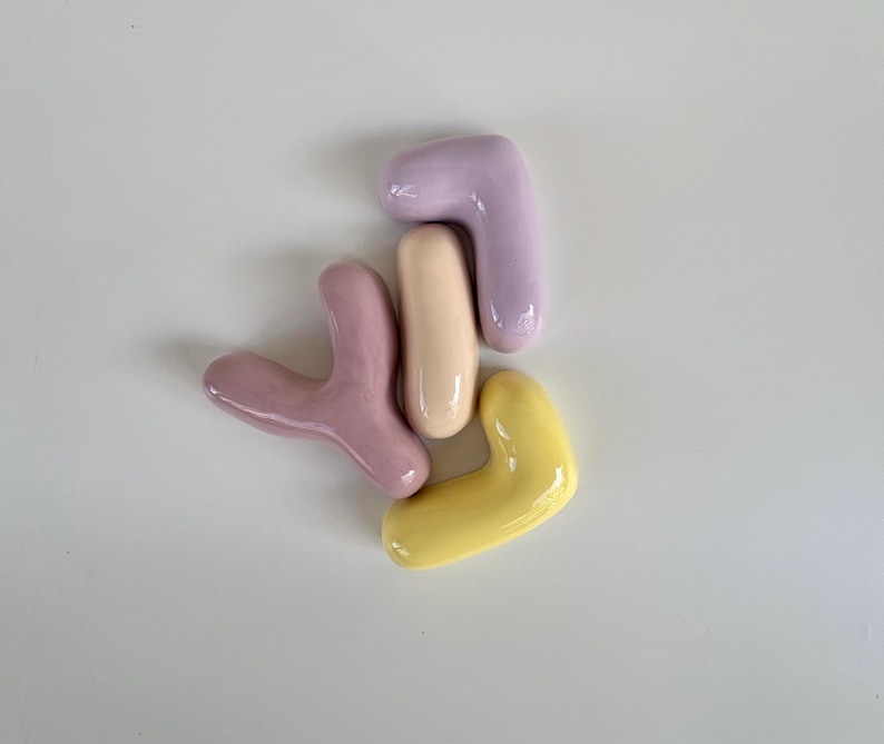 May include: Four abstract ceramic shapes in pastel colours: lavender, yellow, pink, and beige. The shapes are smooth and glossy, with rounded edges. The arrangement is scattered on a white surface.