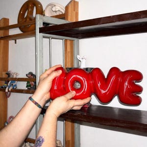 Puede incluir: Una escultura de cerámica roja brillante con la palabra "LOVE". La escultura está siendo sostenida por una persona. Otros objetos decorativos son visibles en estantes en el fondo, incluyendo un pretzel y figuritas.