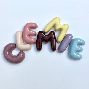 May include: A collection of ceramic letters spelling "LEMMME" in a range of pastel colours. The letters are arranged on a white surface, each with a glossy finish. The colours include shades of purple, pink, brown, yellow, and blue.