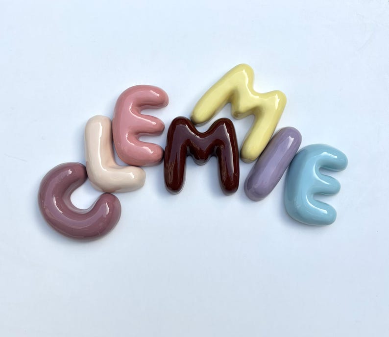 May include: A collection of ceramic letters spelling "LEMMME" in a range of pastel colours. The letters are arranged on a white surface, each with a glossy finish. The colours include shades of purple, pink, brown, yellow, and blue.