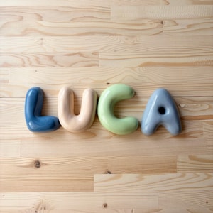 K&ouml;nnte beinhalten: Vier einzelne Keramikbuchstaben, die den Namen LUCA bilden. Der Buchstabe L ist dunkelblau, der Buchstabe U ist blassbeige, der Buchstabe C ist sanft hellgr&uuml;n und der Buchstabe A ist ged&auml;mpft blaugrau. Jeder Buchstabe hat eine glatte, runde Form.