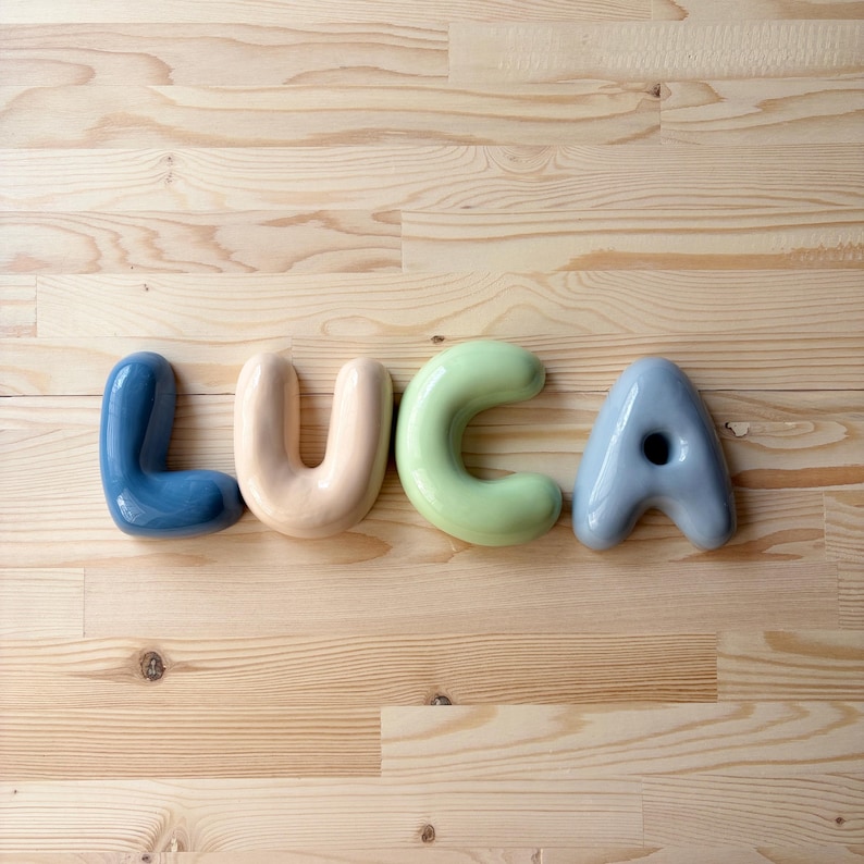 K&ouml;nnte beinhalten: Vier einzelne Keramikbuchstaben, die den Namen LUCA bilden. Der Buchstabe L ist dunkelblau, der Buchstabe U ist blassbeige, der Buchstabe C ist sanft hellgr&uuml;n und der Buchstabe A ist ged&auml;mpft blaugrau. Jeder Buchstabe hat eine glatte, runde Form.