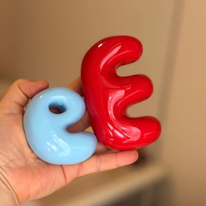 K&ouml;nnte beinhalten: Ein glatter, hellblauer Keramikbuchstabe e mit gl&auml;nzender Oberfl&auml;che und ein gr&ouml;&szlig;erer, glatter, roter Keramikbuchstabe e mit gl&auml;nzender Oberfl&auml;che. Beide Buchstaben haben eine abgerundete, blasenartige Schriftart und eine reflektierende Optik.