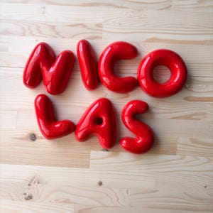 May include: A set of glossy, bright red, three-dimensional letters spelling out the name NICOLAS. Each letter features a smooth, rounded, balloon-like appearance with a reflective surface, arranged in two rows on a light-colored wooden surface.
