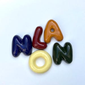 May include: A collection of ceramic letters spelling "Milan". The letters are in a variety of colours: navy blue, burgundy, orange, yellow, and dark green. The letters are arranged on a white background.