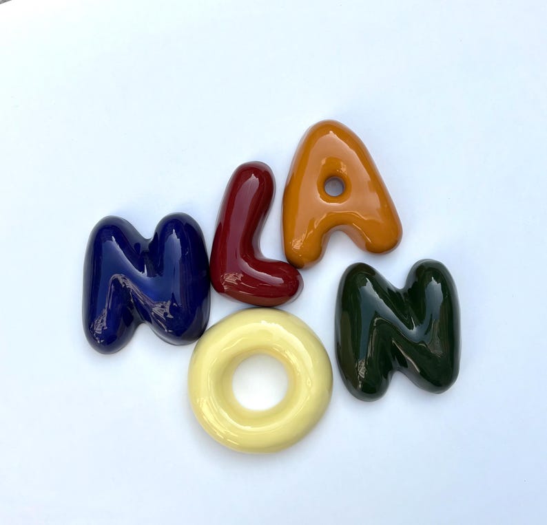 May include: A collection of ceramic letters spelling "Milan". The letters are in a variety of colours: navy blue, burgundy, orange, yellow, and dark green. The letters are arranged on a white background.