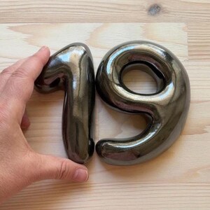 Może przedstawiać: Para tr&oacute;jwymiarowych ceramicznych cyfr, jeden i dziewięć, o gładkim, odblaskowym, metalicznym ciemnoszarym wykończeniu. Cyfry mają zaokrąglony, wypukły wygląd z błyszczącą powierzchnią, kt&oacute;ra chwyta światło, tworząc refleksy na ich zakrzywionych formach.
