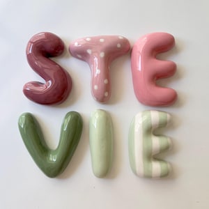 Könnte beinhalten: Sechs einzelne Keramikbuchstaben, die den Namen STEVIE bilden. Das S ist dunkelweinrot, das T ist hellrosa mit weißen Punkten, das E ist einfarbig hellrosa, das V ist olivgrün, das I ist salbeigrün und das E ist salbeigrün mit weißen Streifen.