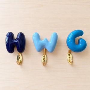Op de afbeelding: Drie wandhaken in de vorm van de letters H, W en G. De H is donker marineblauw, de W is lichtblauw en de G is felblauw. Elke letter heeft een glanzende afwerking en is bevestigd aan een goudkleurige metalen haak met twee schroefgaten.