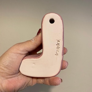 May include: A ceramic letter J in a soft pink hue with a smooth, matte finish. The piece features a small circular hole at the top for hanging and the text "orsayors" is clearly stamped into the lower right side of the letter's surface.