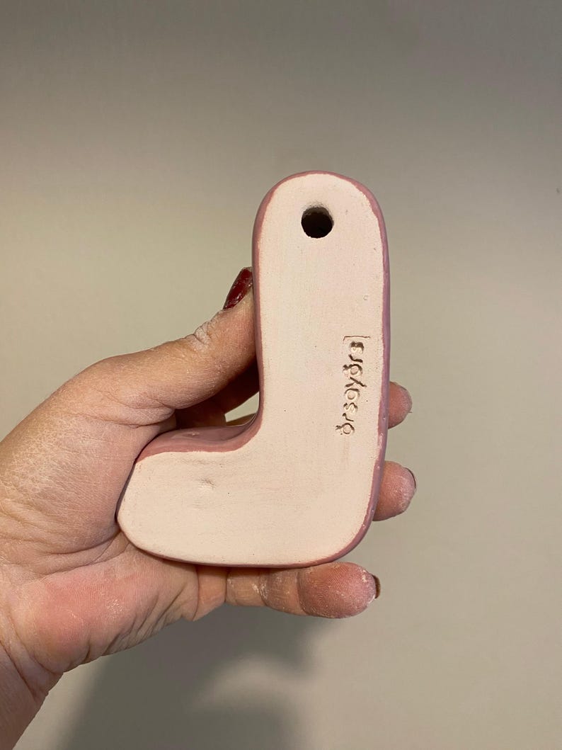 May include: A ceramic letter J in a soft pink hue with a smooth, matte finish. The piece features a small circular hole at the top for hanging and the text "orsayors" is clearly stamped into the lower right side of the letter's surface.