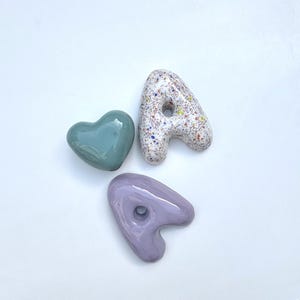 May include: A top-down view of three ceramic pieces: a teal heart, a speckled letter "A", and a lavender letter "A". The letter "A" is white with colourful speckles. The objects have a glossy, smooth finish.