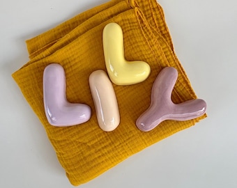 May include: Four ceramic decorative objects in pastel colors, including lavender, yellow, and peach, are arranged on a folded mustard-yellow cloth. The shapes vary, with two L-shaped pieces and two organic forms. The background is white.