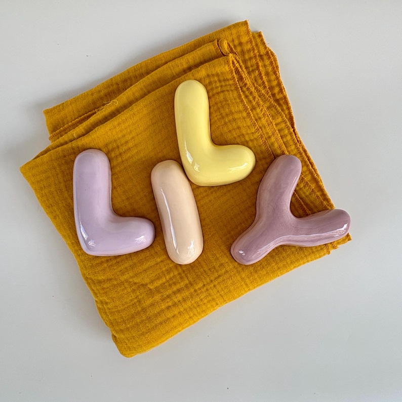 May include: Four ceramic decorative objects in pastel shades, including lavender, yellow, and peach, are arranged on a folded mustard-yellow cloth. The shapes vary, with two L-shaped pieces and two organic forms. The background is white.
