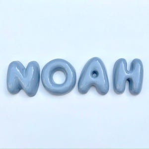 May include: The name "NOAH" is displayed in four light blue, glossy, bubble-style letters. The letters are arranged horizontally against a white background. The letters have a rounded, three-dimensional appearance.