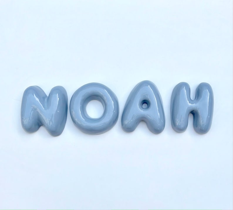 May include: The name "NOAH" is displayed in four light blue, glossy, bubble-style letters. The letters are arranged horizontally against a white background. The letters have a rounded, three-dimensional appearance.