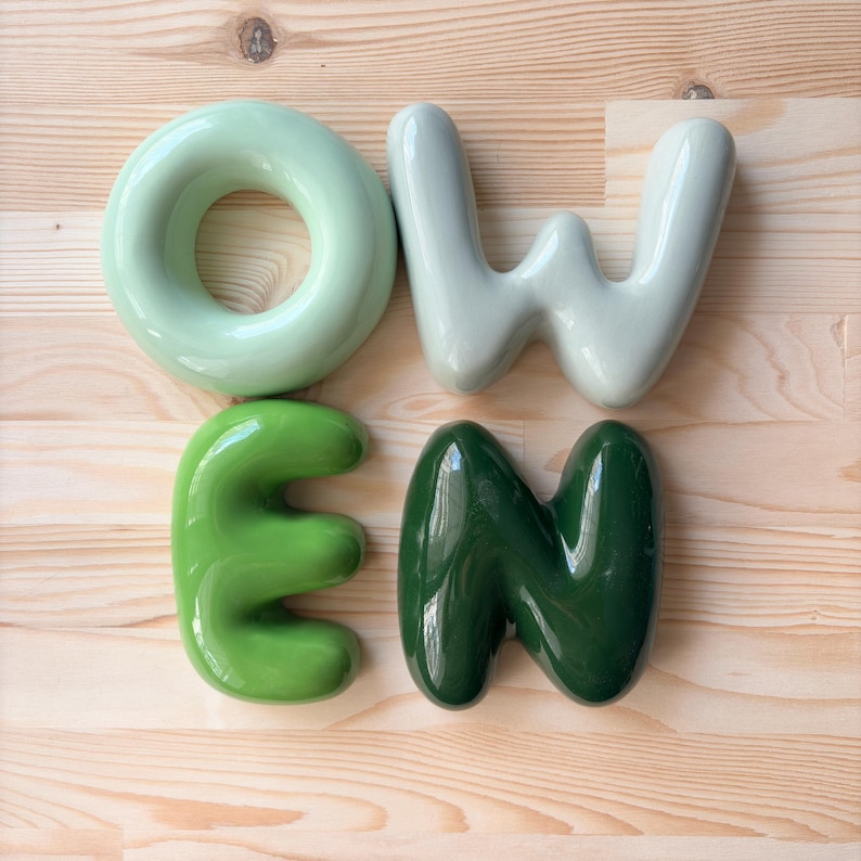 May include: Four glossy, rounded ceramic letters spelling the word "OWEN". The "O" is light mint green, the "W" is pale grey, the "E" is bright lime green, and the "N" is a deep forest green. Each letter has a smooth, inflated, bubble-like appearance.