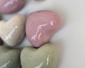 Może przedstawiać: Kolekcja dekoracyjnych przedmiotów w kształcie serca w różnych pastelowych kolorach, w tym różowym, beżowym i zielonym. Błyszczące wykończenie odbija światło, podkreślając gładkie, zaokrąglone kształty. Ułożone na białej powierzchni.