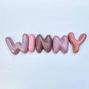 May include: The word "WINNY" spelled out in ceramic letters. The letters are in shades of pink and mauve, with a glossy finish. The letters are arranged horizontally against a white background.