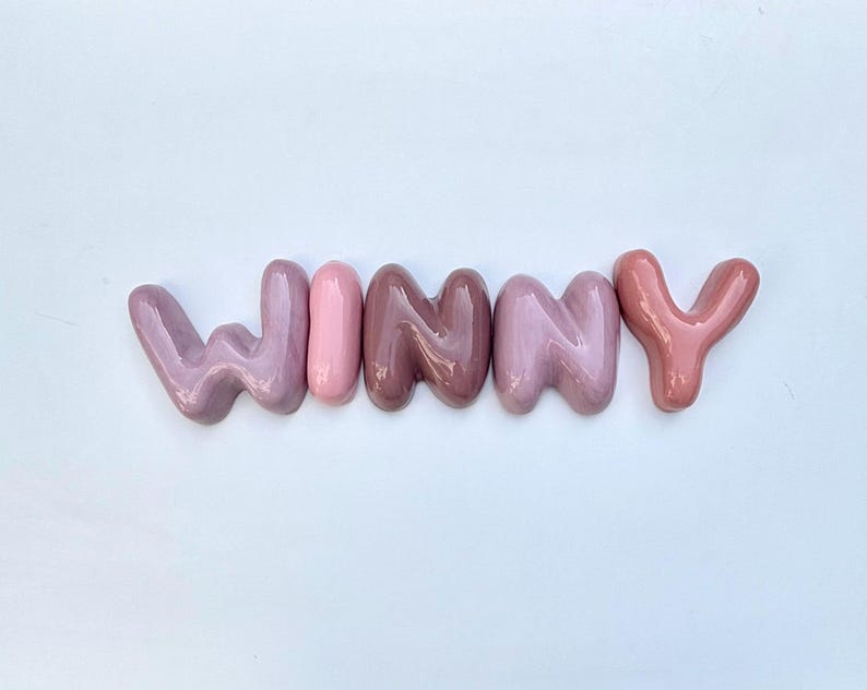 May include: The word "WINNY" spelled out in ceramic letters. The letters are in shades of pink and mauve, with a glossy finish. The letters are arranged horizontally against a white background.