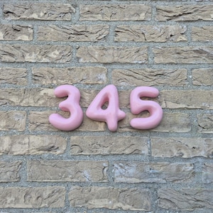 May include: Three-dimensional house numbers 3, 4, and 5 in a soft, rounded, bubble-like font. The numbers are a solid, matte pastel pink colour and are mounted horizontally in a row, displaying the sequence 345 against a textured brick wall.