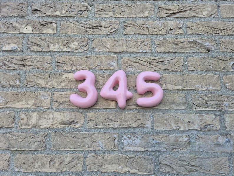 May include: Three-dimensional house numbers 3, 4, and 5 in a soft, rounded, bubble-like font. The numbers are a solid, matte pastel pink colour and are mounted horizontally in a row, displaying the sequence 345 against a textured brick wall.