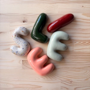 May include: Four ceramic decorative objects shaped like letters or abstract forms. One is white with multicolored speckles, one is dark forest green, one is glossy deep red, one is pale sage green, and one is a soft coral pink, all with smooth surfaces.