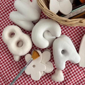May include: Various white ceramic shapes, including a pretzel, a letter "C", a flower, and abstract forms, are arranged on a red and white checked surface. A paintbrush lies nearby, indicating a craft project in progress.