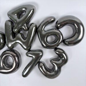 Może przedstawiać: Kolekcja metalicznych, ciemnoszarych cyfr w stylu balonowym, w tym 4, 6, 0, 1 i 3. Te gładkie, zaokrąglone, tr&oacute;jwymiarowe cyfry mają odblaskowe, błyszczące wykończenie i są ułożone w grupę na jednolitym, białym tle.