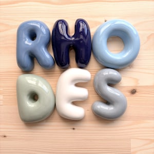Pode incluir: Um conjunto de seis letras de cerâmica brilhante que formam a palavra RHODES. O R é azul claro, o H é azul marinho escuro, o O é azul pálido, o D é verde sálvia, o E é branco sujo e o S é cinza claro. Cada letra tem uma forma lisa e arredondada.
