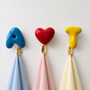 以下が含まれることがあります： ゴールドトーンの金属製ベースが付いた3つの壁掛けフック。それぞれ青い文字A、赤いハート、黄色い文字Tの形をした光沢のあるセラミック製トップが付いています。各フックには、ライトブルー、ペールピンク、クリーム色の折りたたまれたタオルが掛けられています。