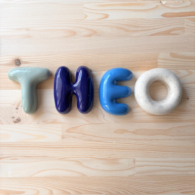 May include: Four individual ceramic letters spelling the name THEO. The letter T is a pale sage green, the H is a deep glossy navy blue, the E is a bright sky blue, and the O is a speckled off-white circular shape with a smooth, rounded finish.