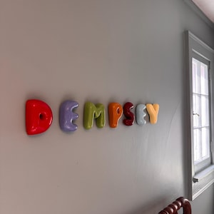May include: A set of wall-mounted letters spelling the name DEMPSEY. Each letter features a smooth, rounded, balloon-like shape in a distinct color, including red, lavender, lime green, orange, dark red, light blue, and yellow.