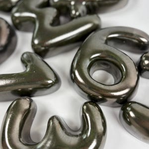 Może przedstawiać: Kolekcja tr&oacute;jwymiarowych, metalicznych ciemnoszarych cyfr o gładkim, błyszczącym wykończeniu. Cyfry, w tym wyraźna sz&oacute;stka i tr&oacute;jka, mają zaokrąglone krawędzie i odblaskową fakturę powierzchni.