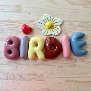 Pode incluir: Um conjunto de letras de cerâmica que formam a palavra BIRDIE em várias cores brilhantes, incluindo malva, lavanda, amarelo, vermelho escuro, rosa e azul. Um pequeno coração vermelho e uma flor branca com pétalas amarelas estão acima.