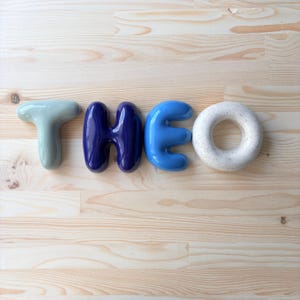 K&ouml;nnte beinhalten: Vier einzelne Keramikbuchstaben, die den Namen THEO bilden. Der Buchstabe T ist blass salbeigr&uuml;n, das H ist tief dunkelblau gl&auml;nzend, das E ist hell himmelblau und das O ist gesprenkelt cremewei&szlig; mit einer glatten, abgerundeten, blasenartigen Form.