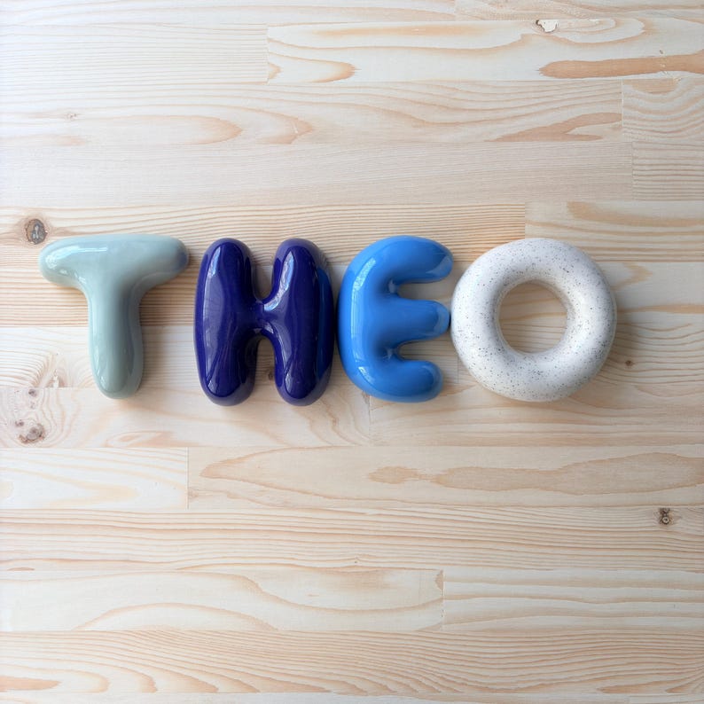 K&ouml;nnte beinhalten: Vier einzelne Keramikbuchstaben, die den Namen THEO bilden. Der Buchstabe T ist blass salbeigr&uuml;n, das H ist tief dunkelblau gl&auml;nzend, das E ist hell himmelblau und das O ist gesprenkelt cremewei&szlig; mit einer glatten, abgerundeten, blasenartigen Form.