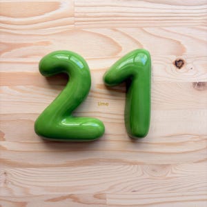 May include: A pair of glossy, lime green ceramic numbers forming the number 21. The digits have a smooth, rounded, three-dimensional shape with a reflective surface. The word lime is printed in small yellow letters between the two digits.