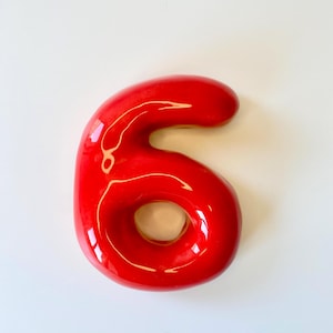 May include: A glossy, bright red ceramic number six with a smooth, rounded, and inflated appearance. The digit features a reflective surface with visible light highlights, creating a three-dimensional effect. The shape is continuous and solid.