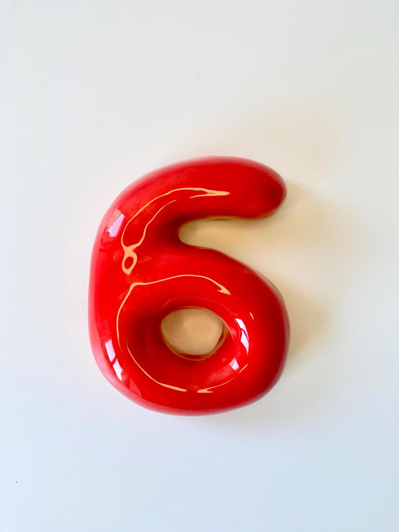 May include: A glossy, bright red ceramic number six with a smooth, rounded, and inflated appearance. The digit features a reflective surface with visible light highlights, creating a three-dimensional effect. The shape is continuous and solid.