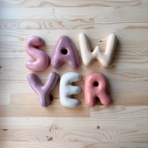 May include: A set of six ceramic letters spelling the word SAWYER. The letters are rounded and puffy in shape, featuring a variety of colors including dusty pink, mauve, cream, and a white speckled finish, with some letters containing holes.
