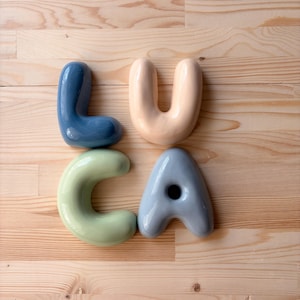 May include: Four individual ceramic letters spelling the name LUCA. The letter L is dark blue, the letter U is a pale peach, the letter C is a soft sage green, and the letter A is a cool grey. Each letter has a smooth, rounded, and glossy finish.